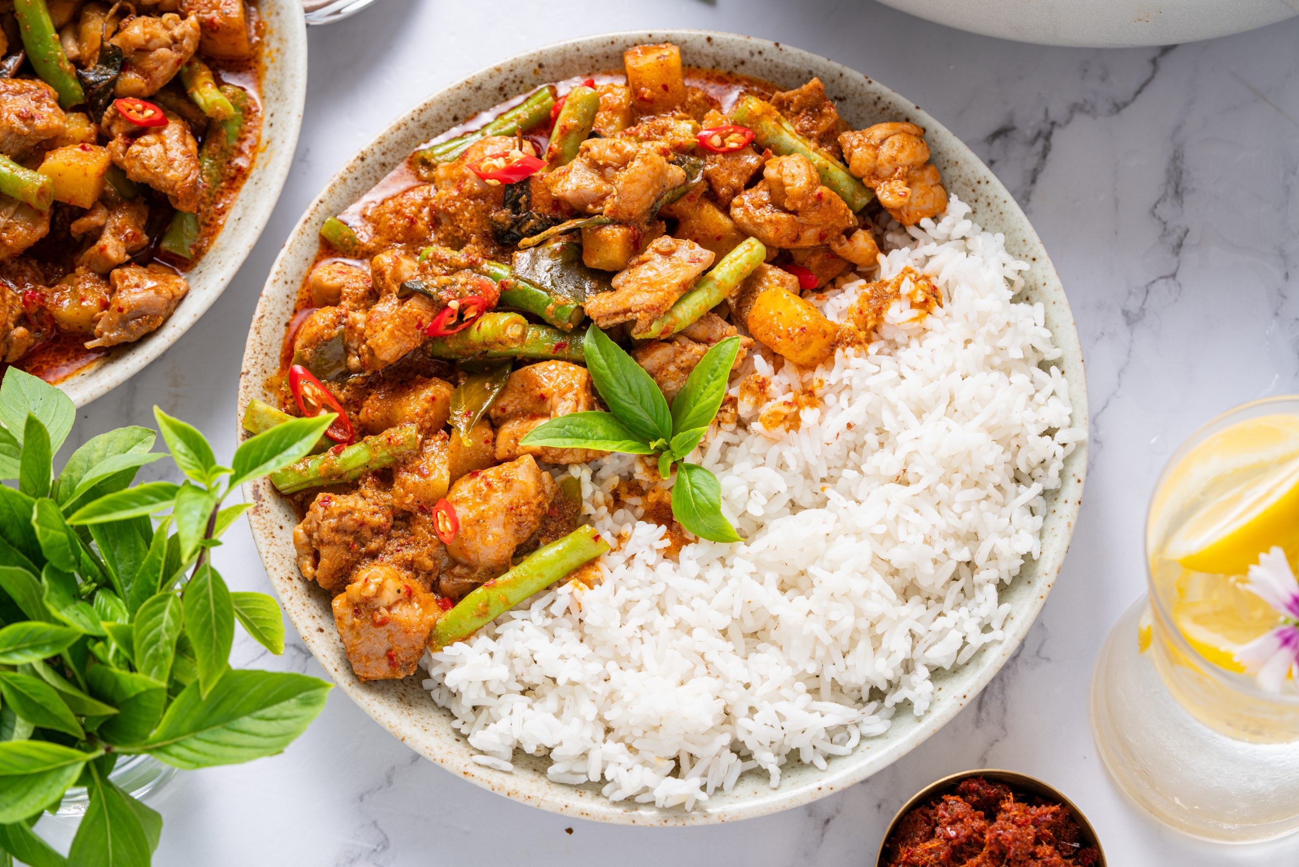Thai chicken Panang curry plated with rice, vibrant red curry sauce with tender chicken, green beans, and basil leaves on a light marble background.