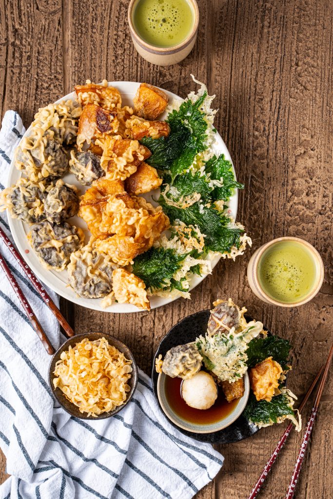 Japanese vegetable tempura platter with mushrooms, sliced pumpkin, and shiso leaves, served with dipping sauce on rustic wooden table.