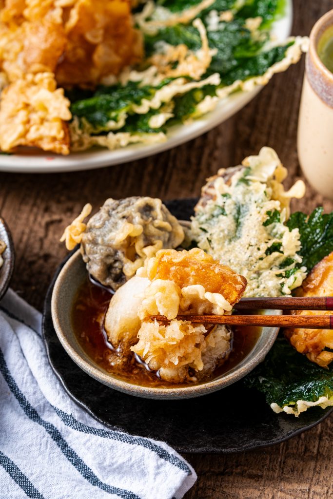 Chopstick holding vegetable tempura dipped in soy-based sauce with grated daikon, showcasing crispy Japanese tempura texture.