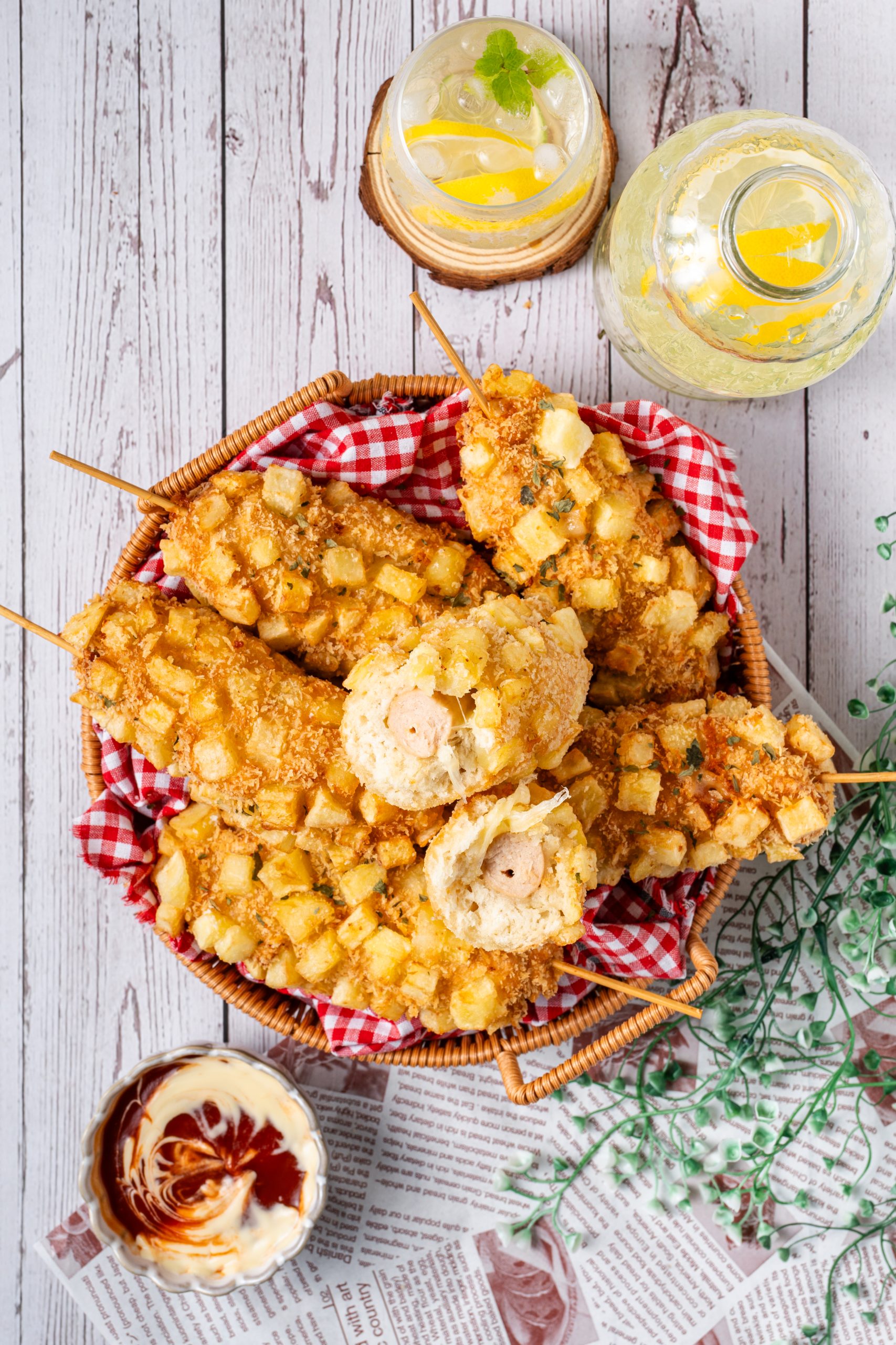 Korean corn dogs coated with crispy diced potatoes, served in a basket with dipping sauce and refreshing lemon drink on a rustic table.