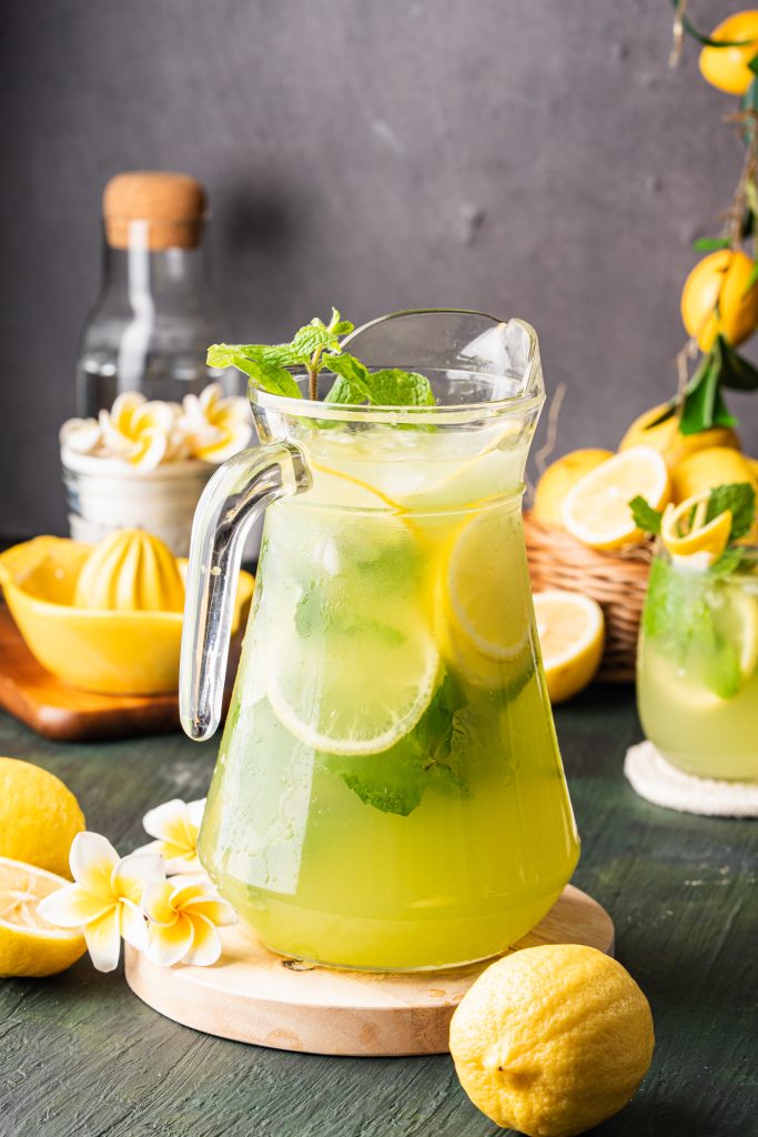 Pitcher of refreshing cucumber lemonade with lemon slices, mint, and ice, styled with fresh lemons and flowers.