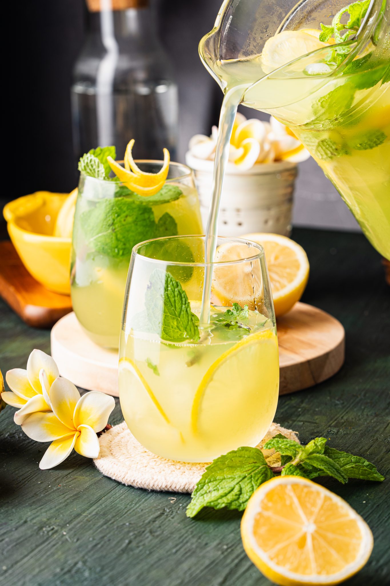 Cucumber lemonade being poured from a glass pitcher into a cup with lemon slices, mint, and ice.