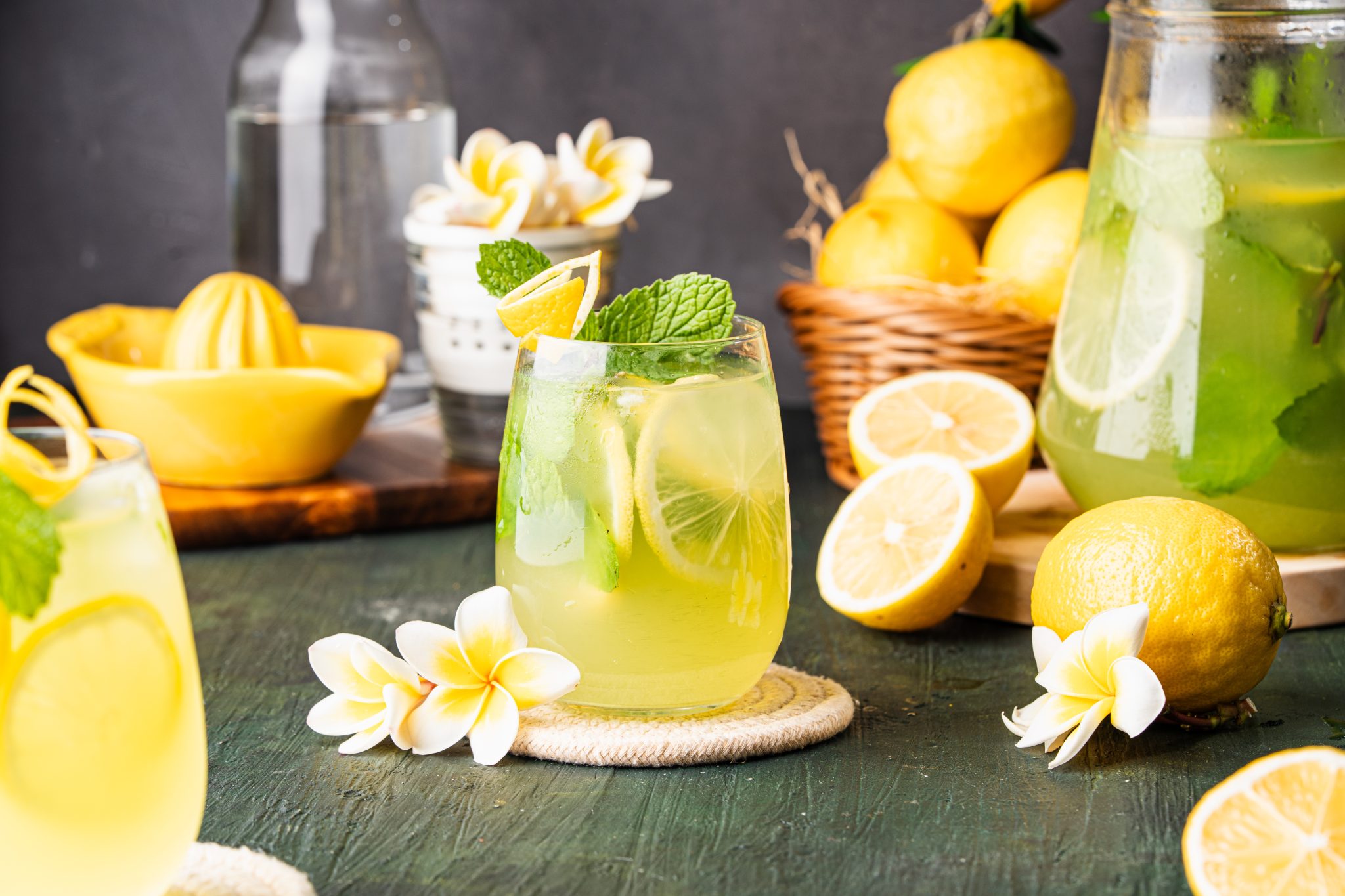 Glass of cucumber lemonade with lemon slices and mint, surrounded by fresh lemons, flowers, and a pitcher.