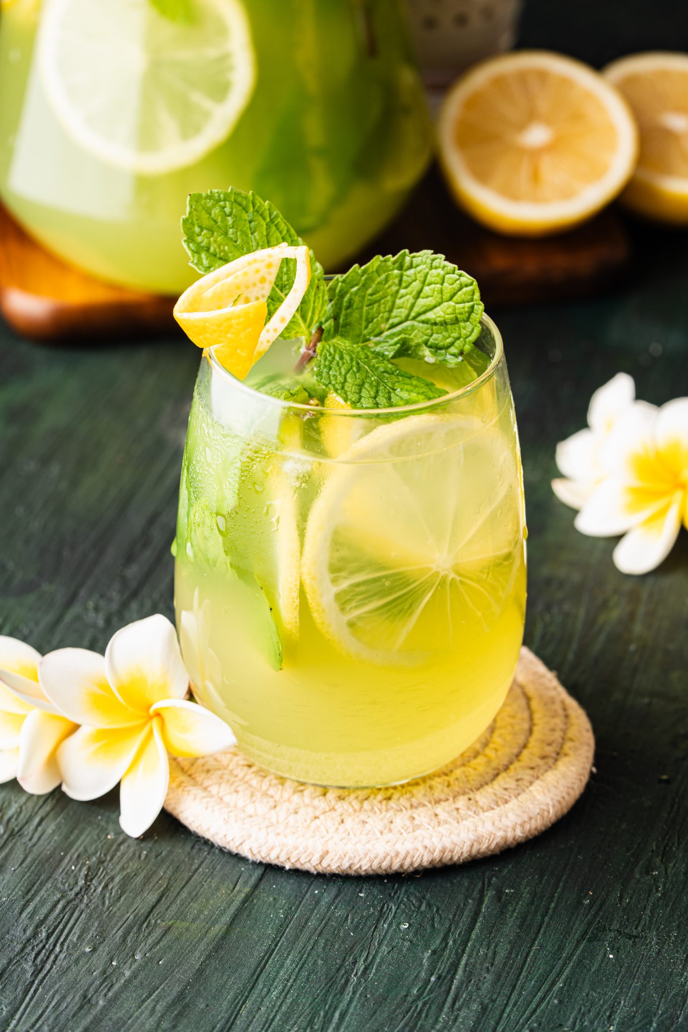 Close-up of cucumber lemonade garnished with lemon peel and fresh mint beside sliced lemons and flowers.