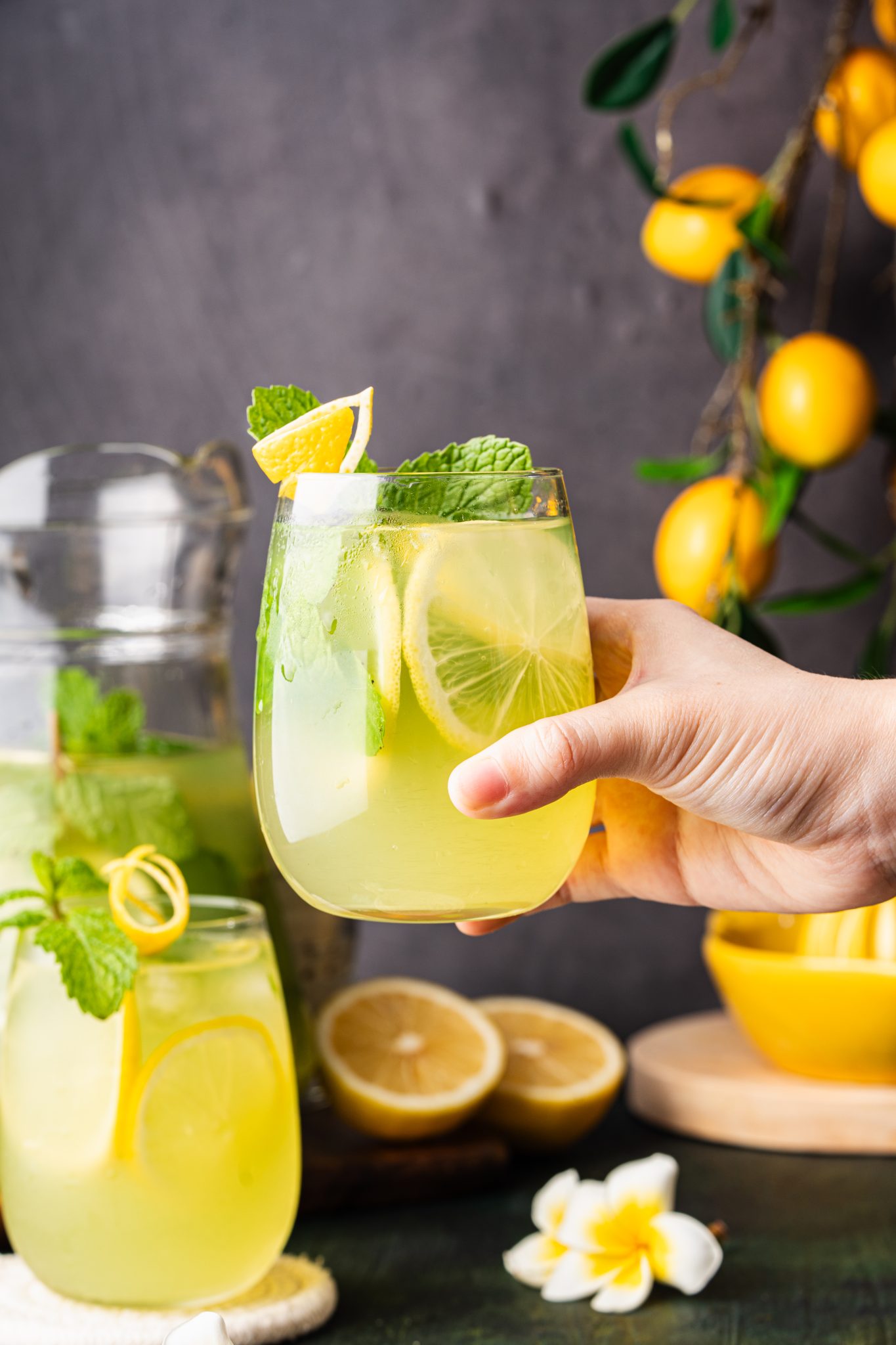 Hand holding a glass of cucumber lemonade with lemon slices, mint, and ice beside a pitcher and fresh lemons.