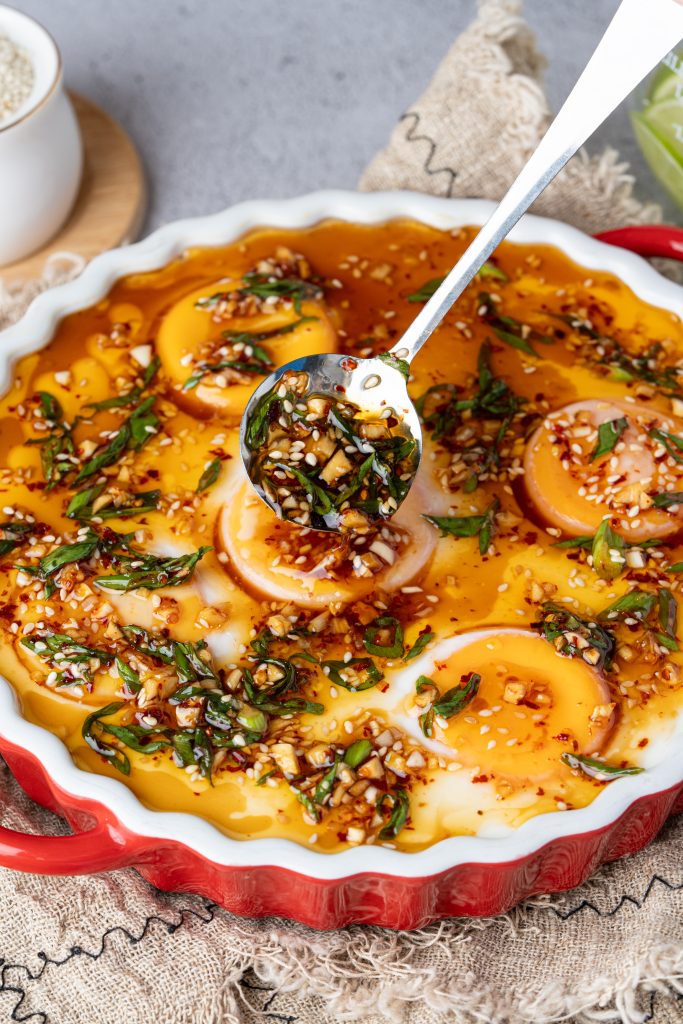 Spoon drizzling chili garlic oil over silky Chinese double steamed eggs topped with scallions and sesame seeds in a red baking dish.