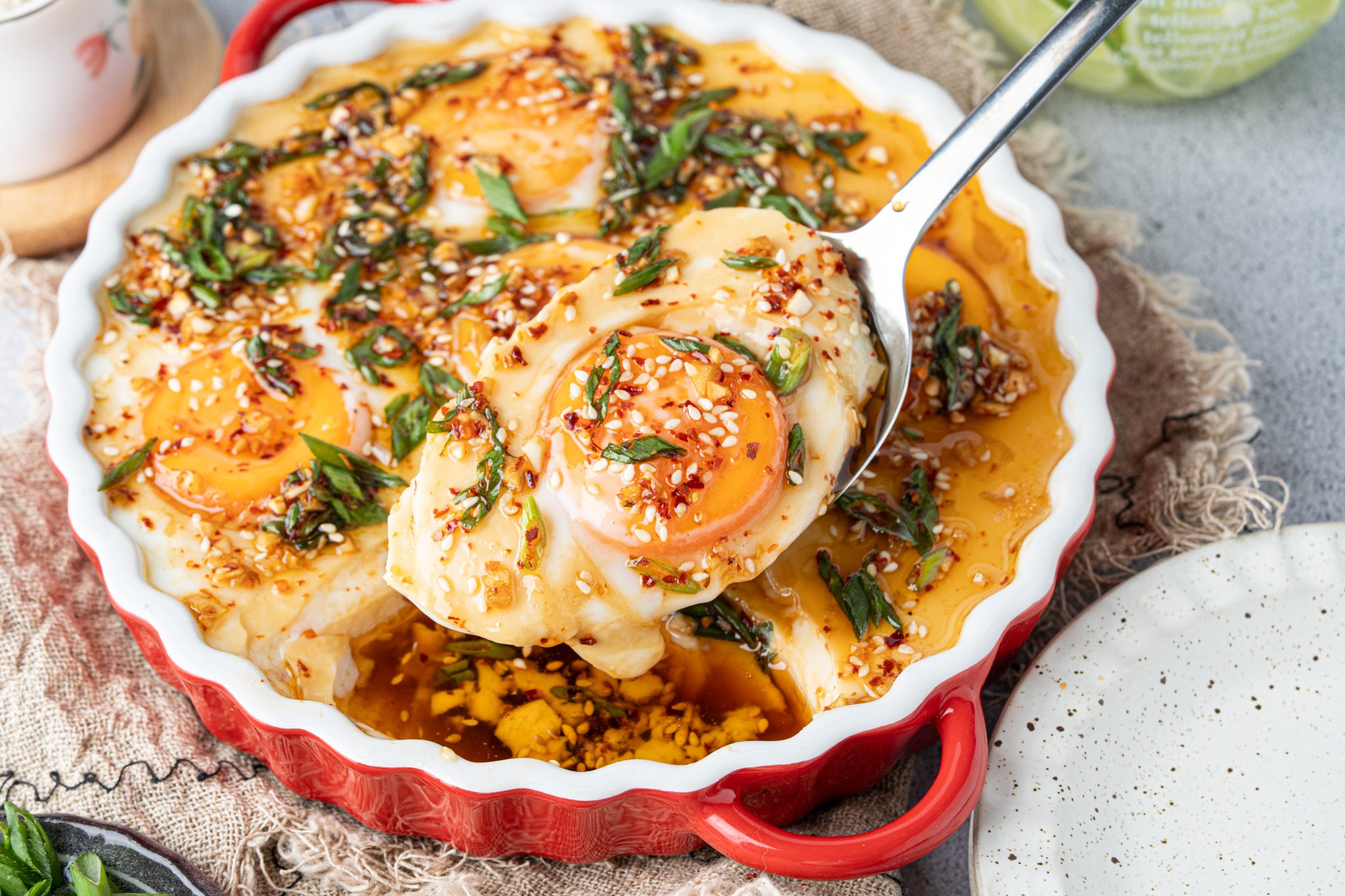 Creamy Chinese steamed egg custard being served from a red baking dish with chili oil, garlic, scallions, and sesame seeds.