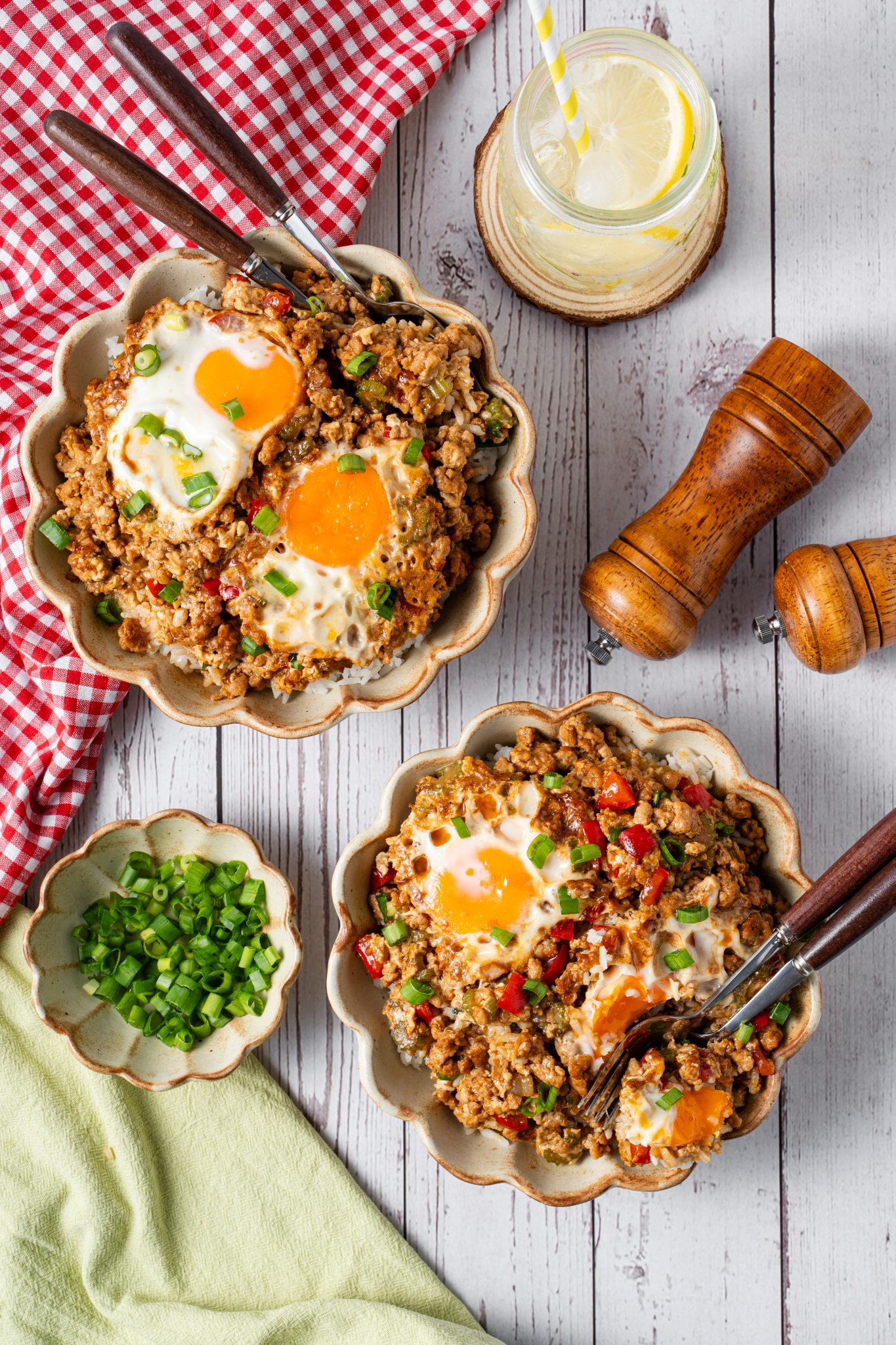 Overhead view of minced meat and fried egg rice bowls with scallions, lemon drink, and gingham cloth.